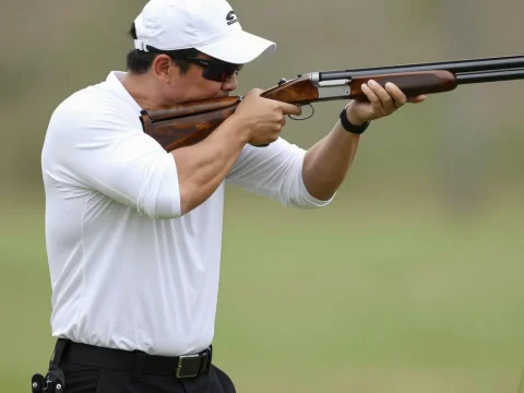 Man Aiming Shotgun Outdoors in White Shirt and Cap